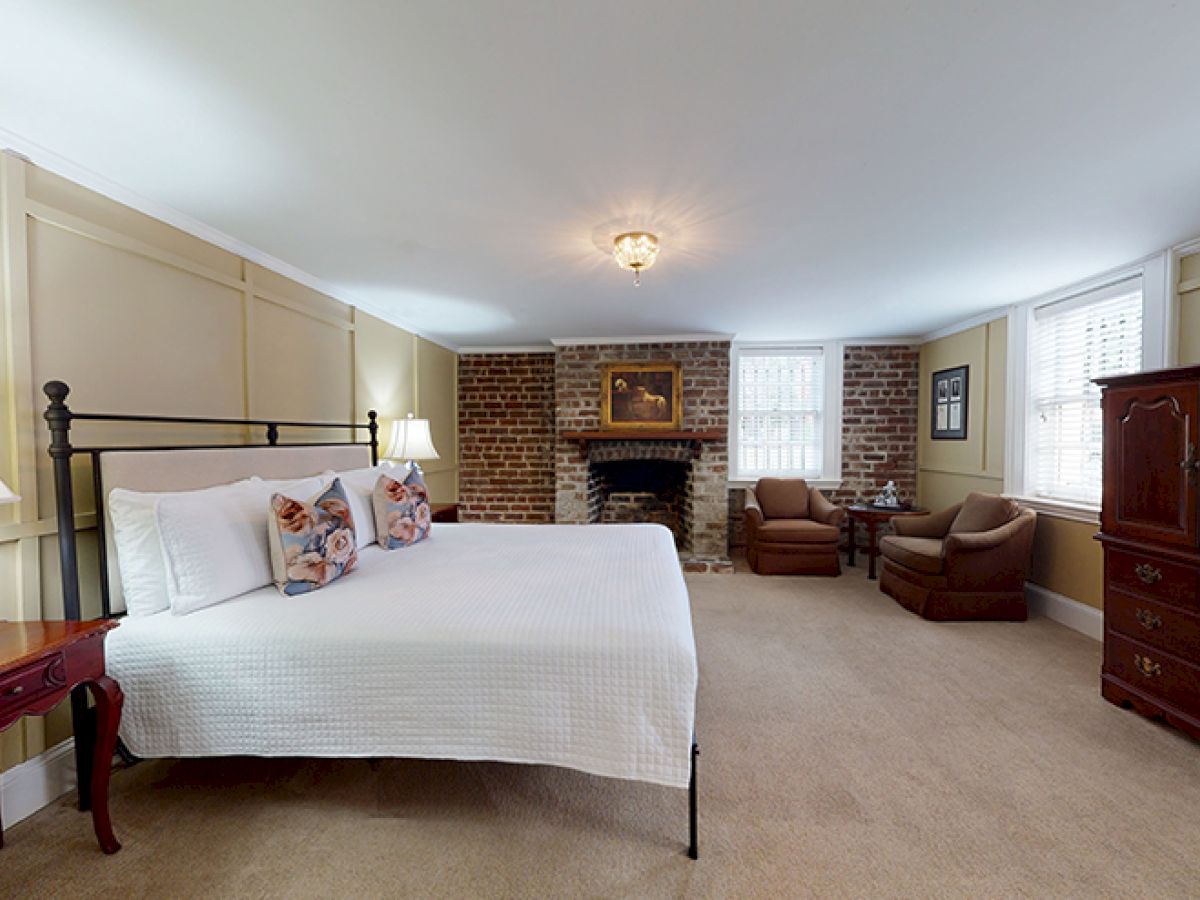 A cozy bedroom with a king-sized bed, brick fireplace, two armchairs, wooden armoire, windows, and bedside tables with lamps.