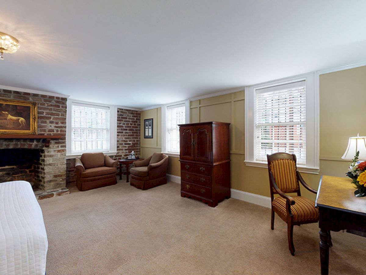 A spacious bedroom features a bed, brick fireplace, two armchairs, a wooden cabinet, a desk with a lamp and flowers, and two windows with blinds.