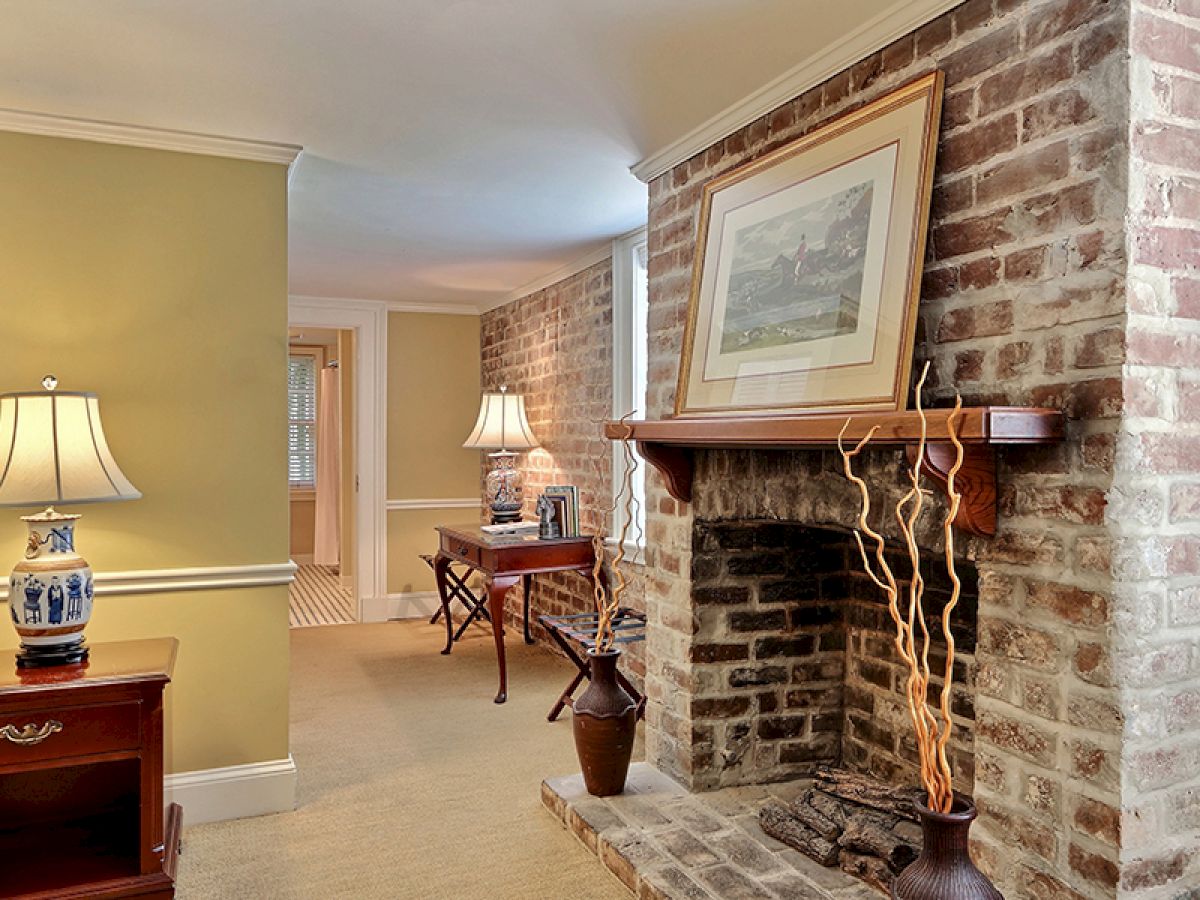 The image shows a cozy living room with a brick fireplace, framed artwork above, tall decorative branches, lamps on tables, and warm lighting.