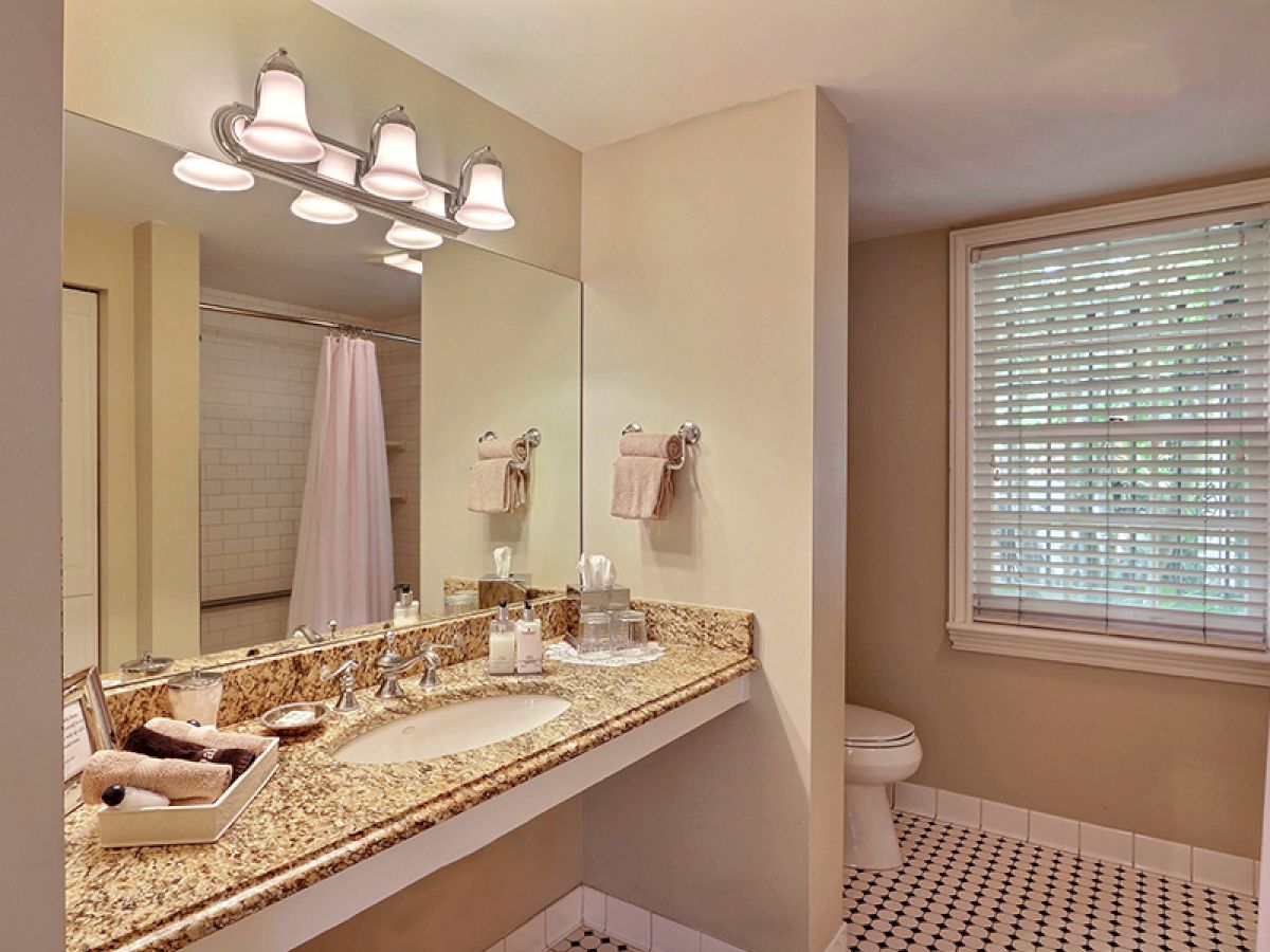 A clean bathroom with a granite countertop, double sink, large mirror, and light fixtures. A toilet and a window with blinds are visible.