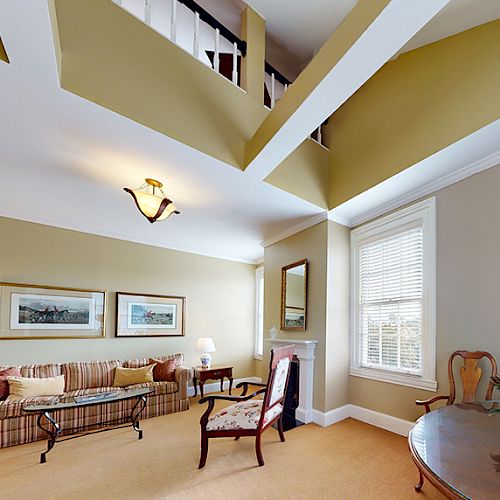 A spacious living room with beige walls, a striped sofa, chairs, a glass coffee table, framed pictures, and a ceiling with exposed beams.