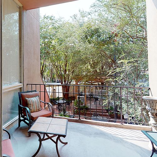 A cozy balcony with seating, a table, and a view of lush green trees and other balconies in the distance.