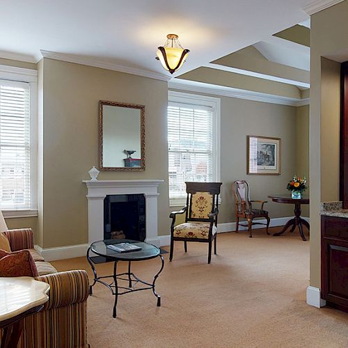 This image shows a cozy living room with a sofa, a fireplace, a coffee table, and a small dining area near a window with blinds.