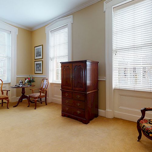 The image shows a room with beige walls, a carpeted floor, a table with two chairs, a wooden cabinet, and a floral armchair near two windows.