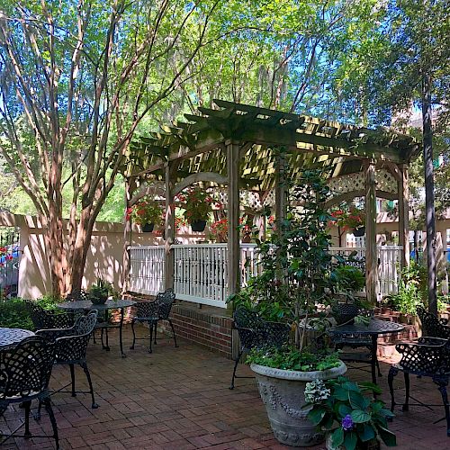 An outdoor patio area with metal chairs and tables, surrounded by trees, potted plants, and a pergola with hanging baskets, under a sunny sky.