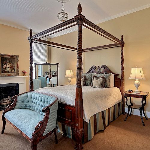 A cozy bedroom features a four-poster bed, blue loveseat, fireplace, lamps, mirrors, and artwork, creating a warm, welcoming ambiance.