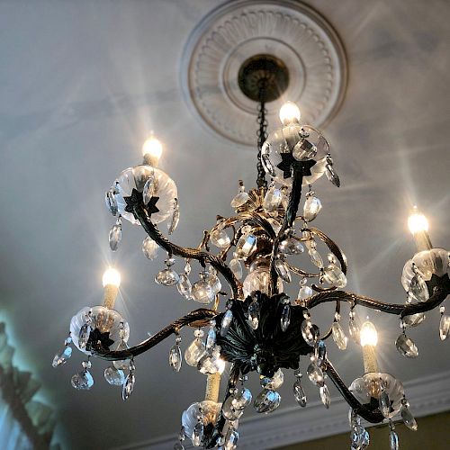 An ornate chandelier with lit candles and crystal decorations hangs from a ceiling with decorative molding, casting light and shadows in the room.