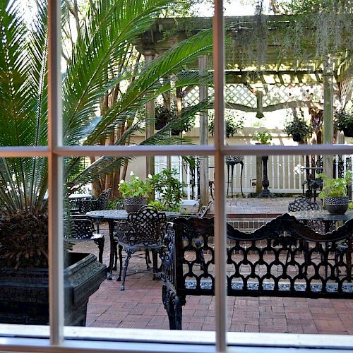 A view through a window of a garden with wrought iron furniture, potted plants, and a pergola with greenery in the background.