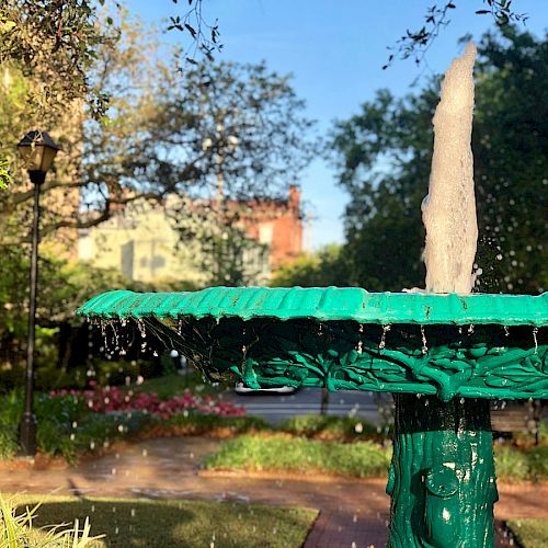 A green fountain in a garden with water spraying upward, surrounded by trees, plants, and pathways on a sunny day.