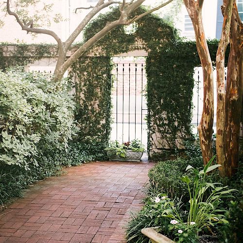 A brick pathway curves along a lush garden with trees, hedges, and a potted plant, leading to a white gate covered in ivy.