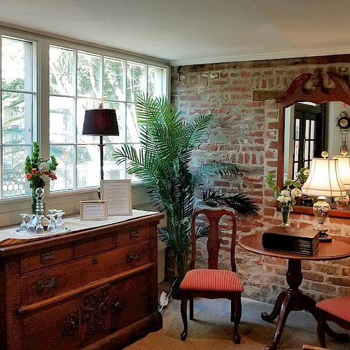 A cozy room with a brick wall, a dresser, a mirror, two chairs, a small table, and multiple lamps, with large windows letting in natural light.