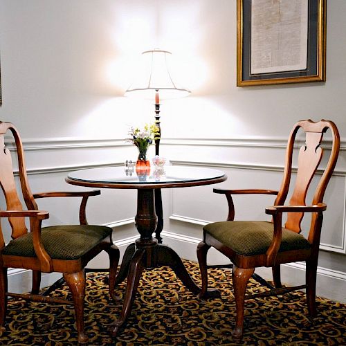 A small round table with two wooden chairs, a lamp, and framed wall art in a cozy, carpeted room with wainscoting on the walls.