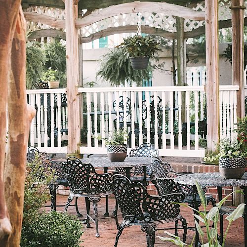 The image shows an outdoor patio area with wrought-iron chairs and tables, surrounded by lush greenery and shaded by a wooden pergola.