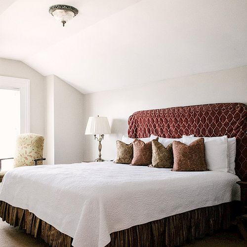 A bedroom with a large bed, white bedding, and several throw pillows. There's a patterned headboard, a window, a lamp, and an armchair in the corner.