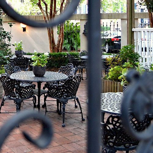 The image shows a charming outdoor patio with wrought iron tables and chairs, surrounded by lush greenery and enclosed by a decorative fence.