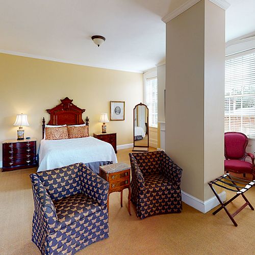 A spacious bedroom with a double bed, two patterned chairs, a small table, a floor mirror, a chest of drawers, and a window with blinds.