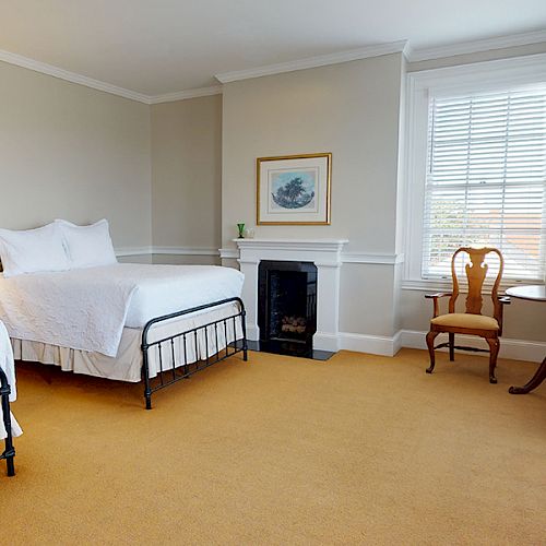 A bedroom with two beds, a fireplace, a table with chairs near the window, a lamp, and a dresser; the room has neutral tones.