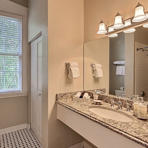 A well-lit bathroom with a large mirror, granite countertop, sink, towels, and shower area with white tiles. There's a window with blinds.