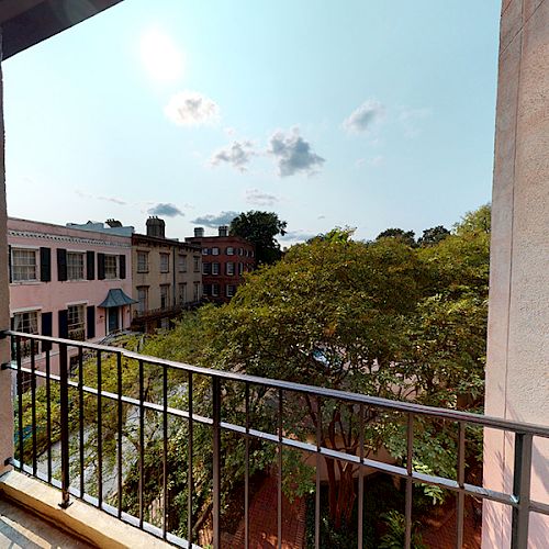 A view from a balcony shows buildings, trees, and a partly cloudy sky. The balcony has a black railing and the walls are a light color.