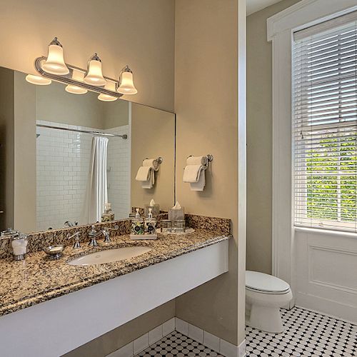 A bathroom with a granite countertop, a large mirror, under-sink cabinets, wall lighting, a window with blinds, and a toilet in the corner.