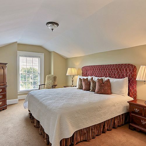 A cozy bedroom with a large bed, two bedside tables with lamps, a wooden dresser, and a window with blinds, featuring warm and inviting decor.