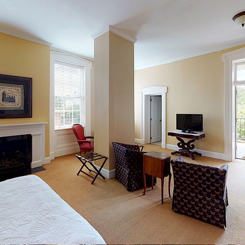 A cozy room with a bed, fireplace, TV, chairs, a small desk, and access to a balcony through a glass door, with natural light streaming in.