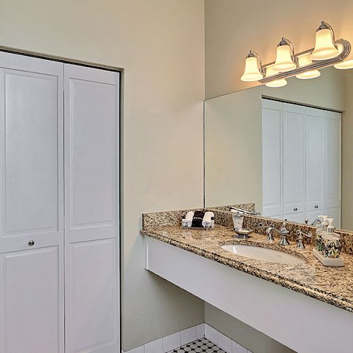 A bathroom with a large mirror, granite countertop, light fixtures above the mirror, and white cabinets. Additional room visible in the mirror.