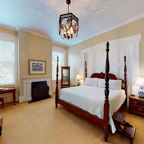 A cozy bedroom with a four-post bed, nightstands with lamps, a fireplace, a wooden desk, a chair, and large windows providing natural light.