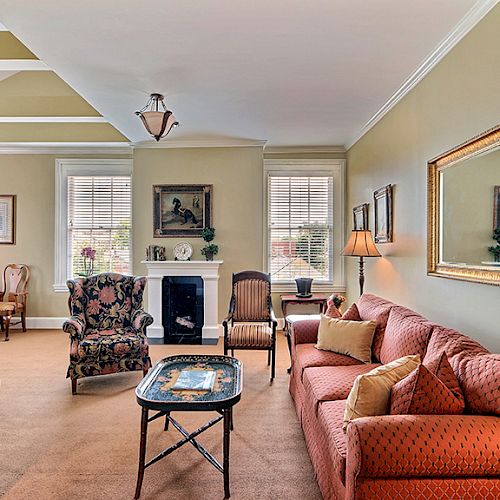 A well-lit living room with a red sofa, patterned armchair, coffee table, large mirror, fireplace, and windows with shutters and wall art.