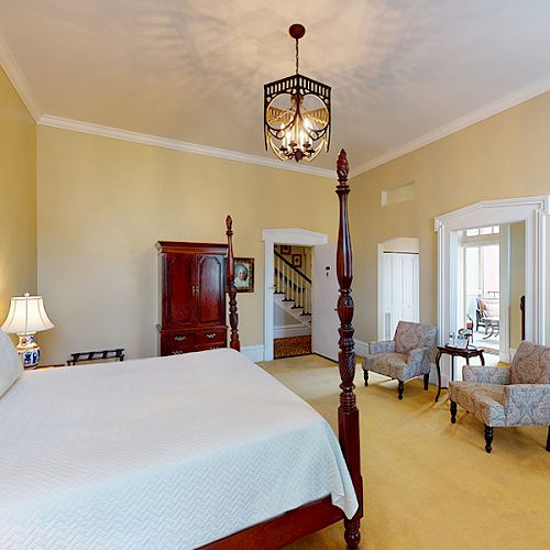 This image shows a spacious bedroom with a large bed, classic furniture, chairs, a chandelier, and access to another room.