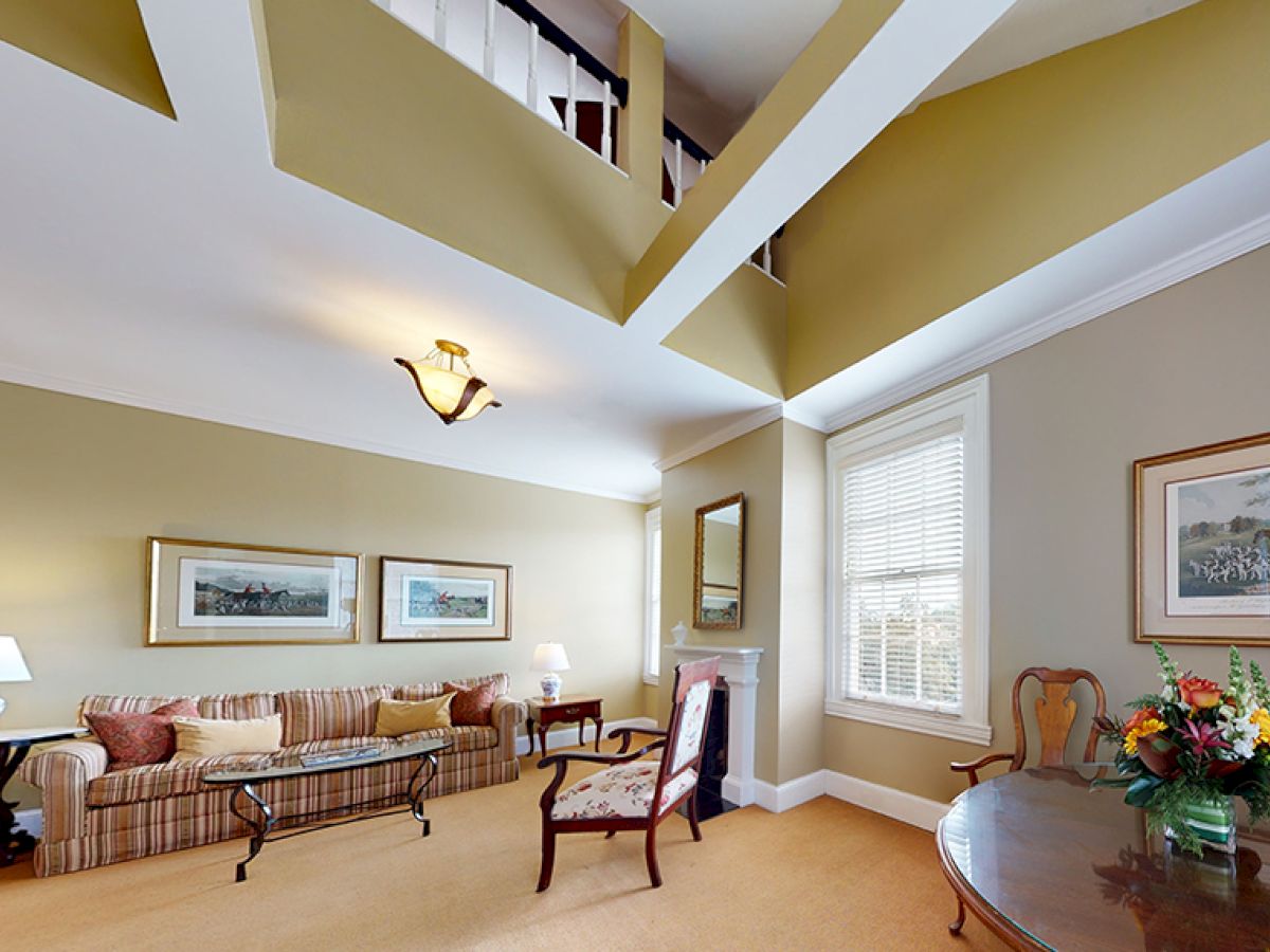 This image shows a living room with a high ceiling, striped sofa, armchair, coffee table, artwork on walls, a window, and a round table with flowers.