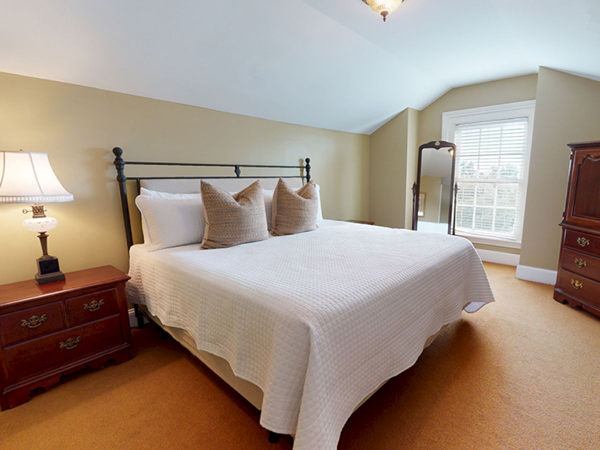 A cozy bedroom features a bed with white linens and two pillows, nightstands with a lamp, an armoire, and a window with shutters.