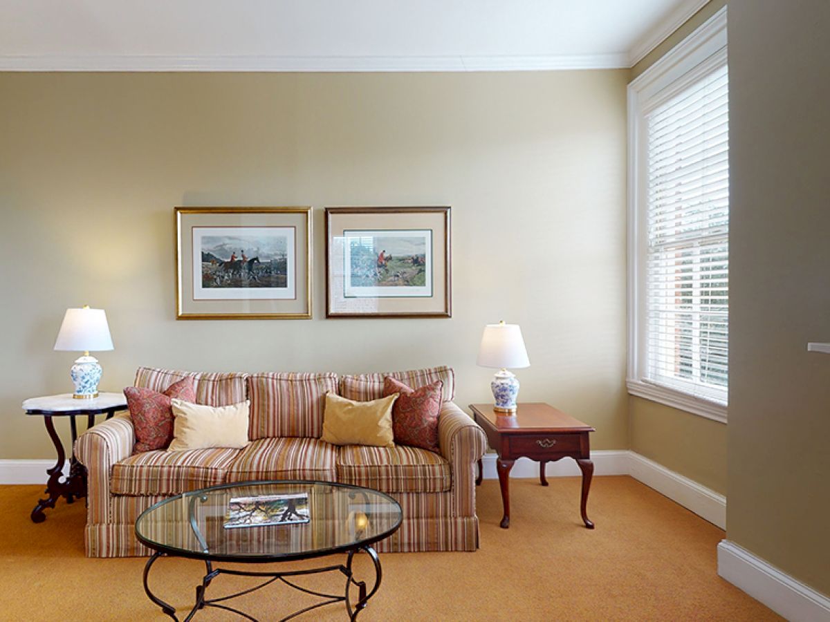 A cozy living room featuring a striped sofa with cushions, two side tables with lamps, framed artworks, and a glass coffee table.