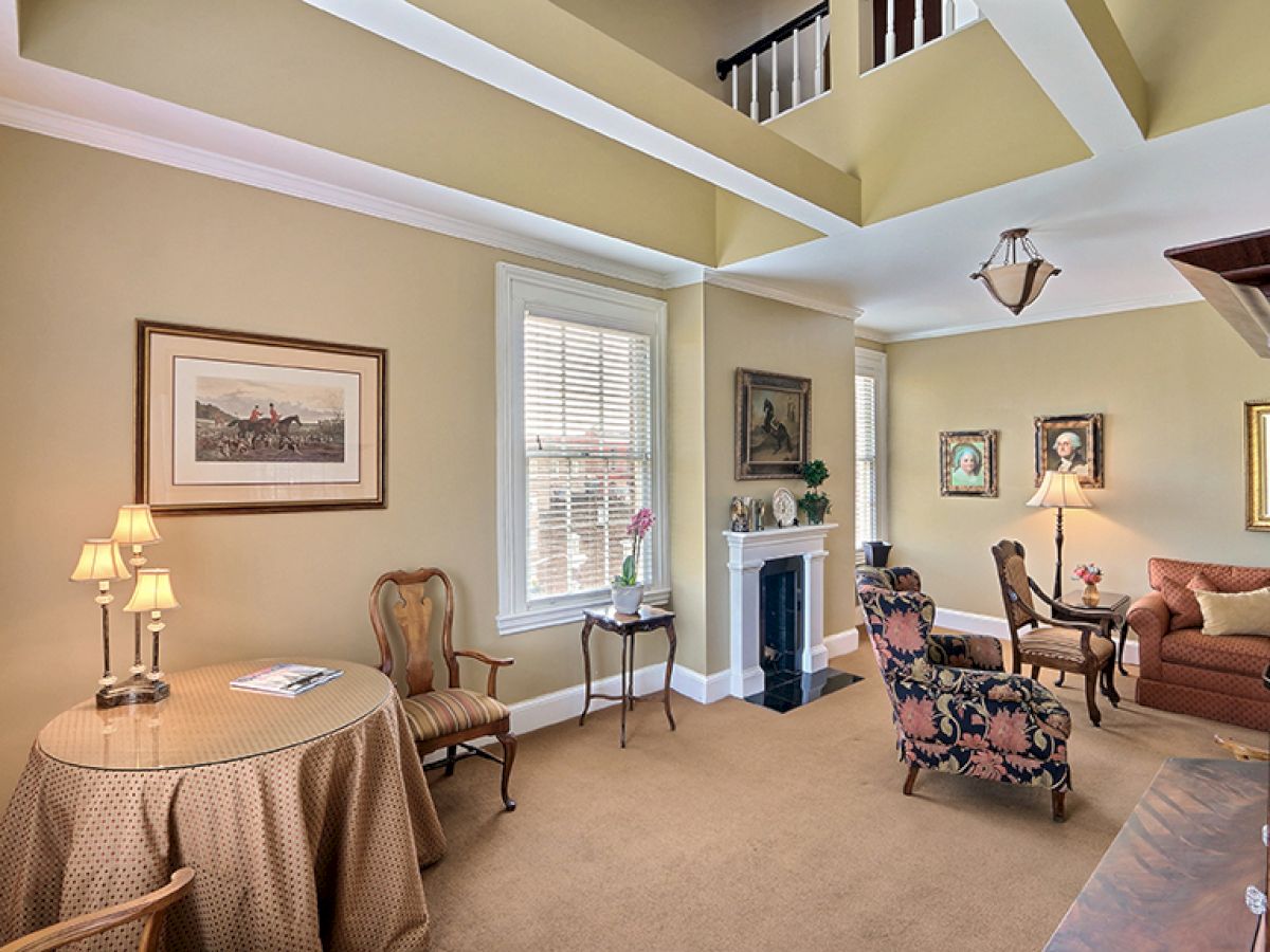 A cozy living room with beige walls, several seating options, framed artwork, a table with a lamp, and a fireplace, under a high ceiling.