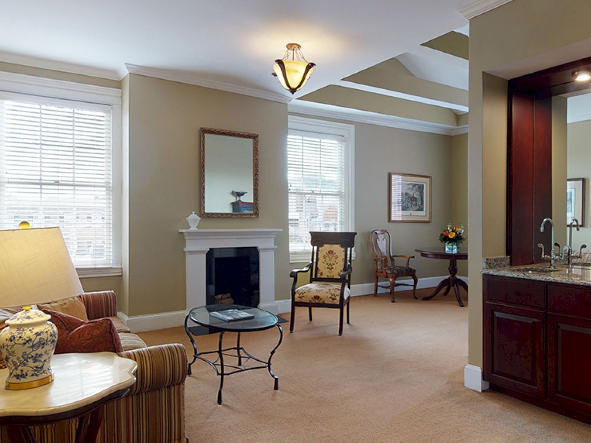 A cozy living room with a sofa, coffee table, lamp, chairs, fireplace, and large windows is shown, adjacent to a small kitchen area.