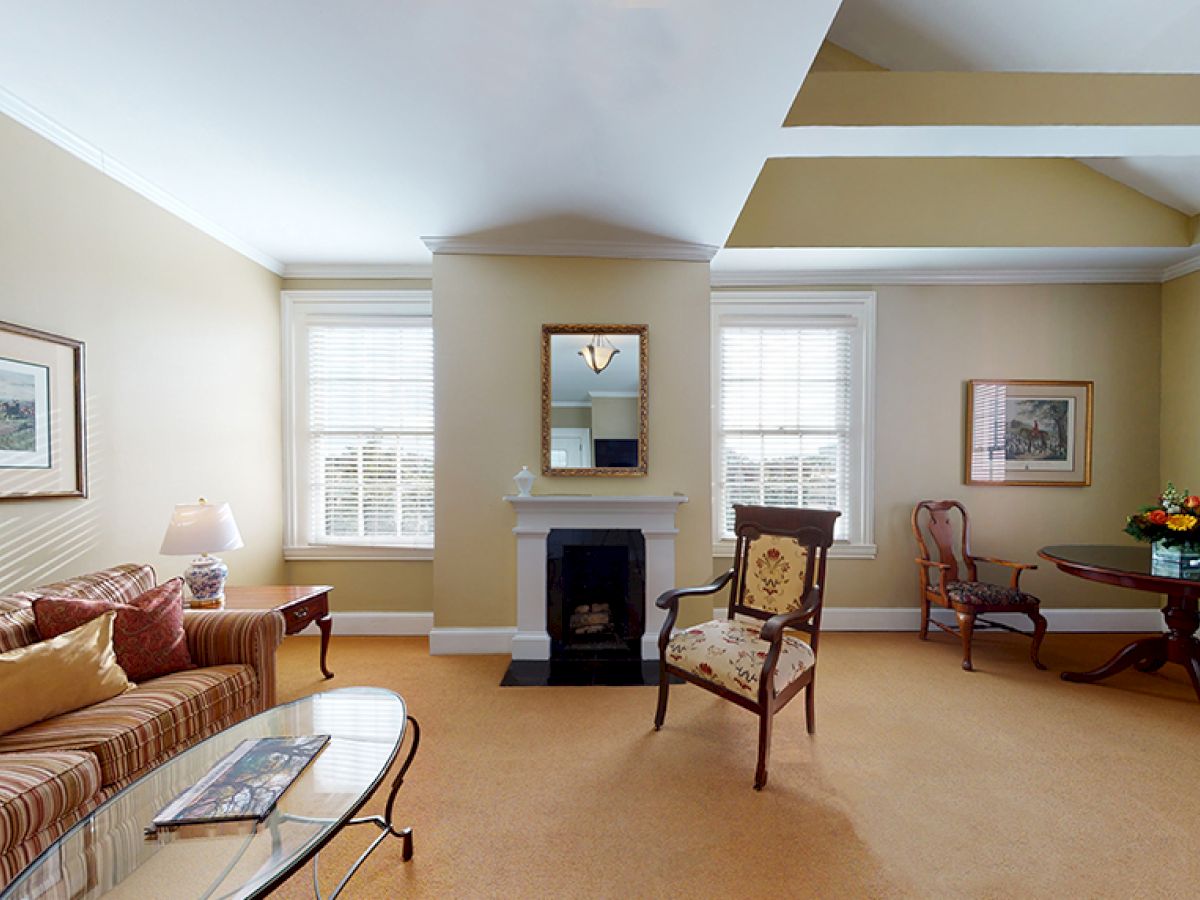 A living room with beige walls featuring a striped sofa, a glass coffee table, a fireplace, chairs, a round table with flowers, and wall art.