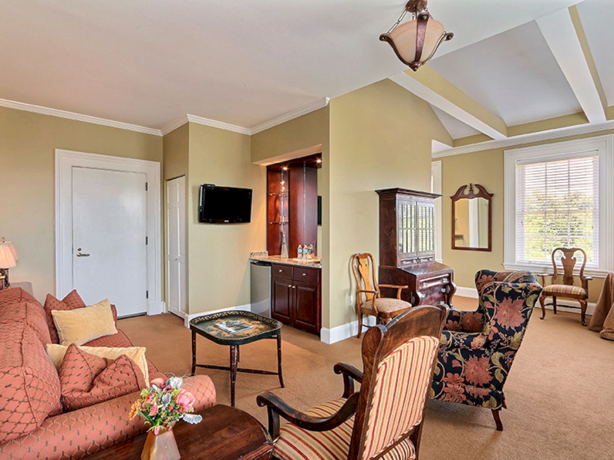 A living room with elegant furniture including sofas, chairs, and a small table. There’s a wall-mounted TV, a side table with a mirror, and a window.