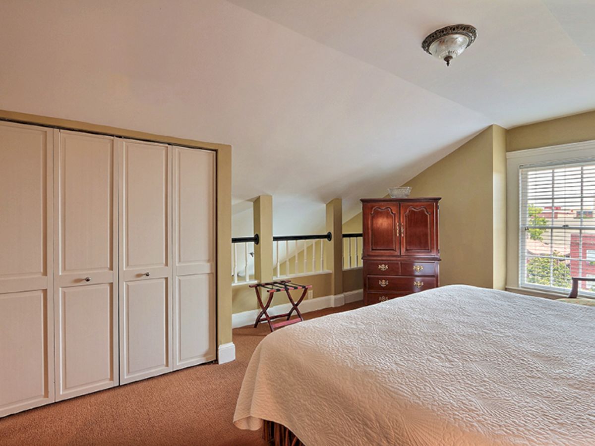 A cozy bedroom with a bed, wooden dresser, closed wardrobe, window with blinds, and a luggage stand against light-colored walls and carpet.