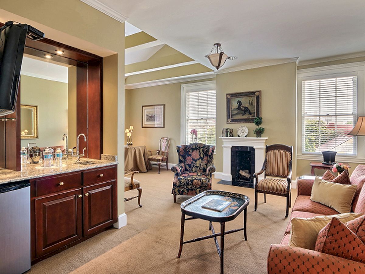 The image shows a well-furnished living room with a sofa, armchairs, a TV, a fireplace, a small kitchenette area, and decorative elements.