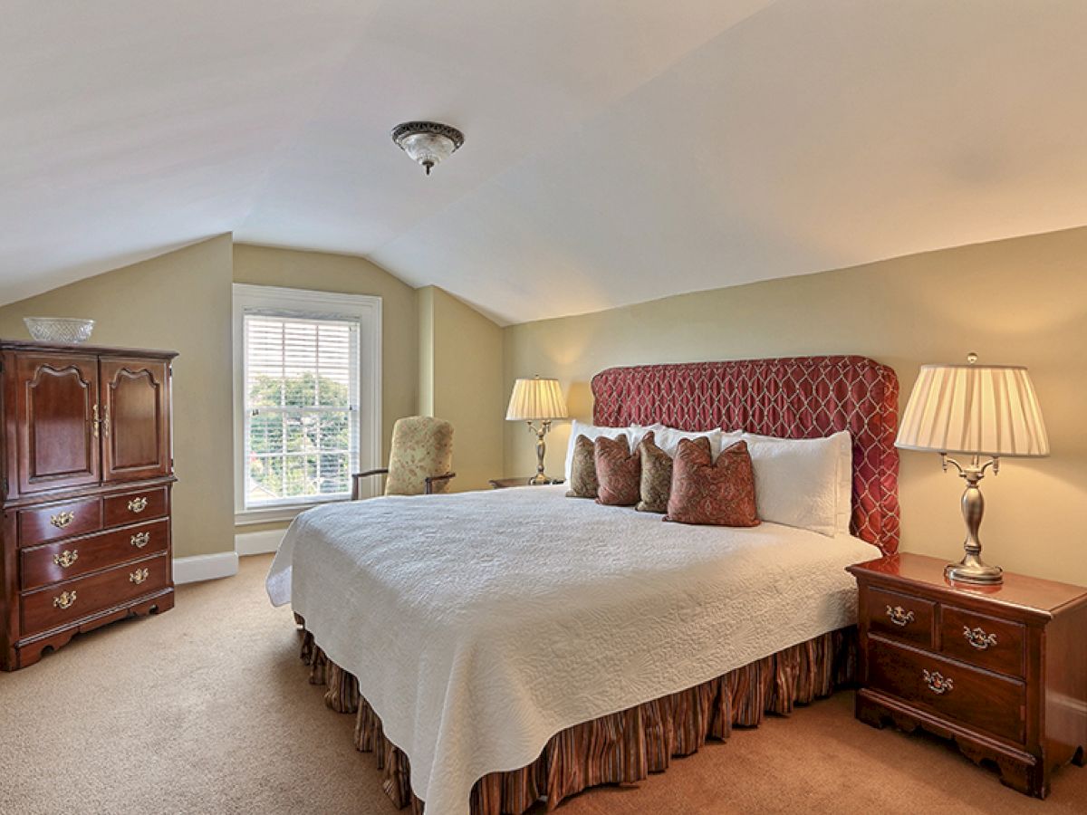 A spacious bedroom with a large bed, patterned headboard, two bedside tables with lamps, a wooden wardrobe, and a window with shutters.