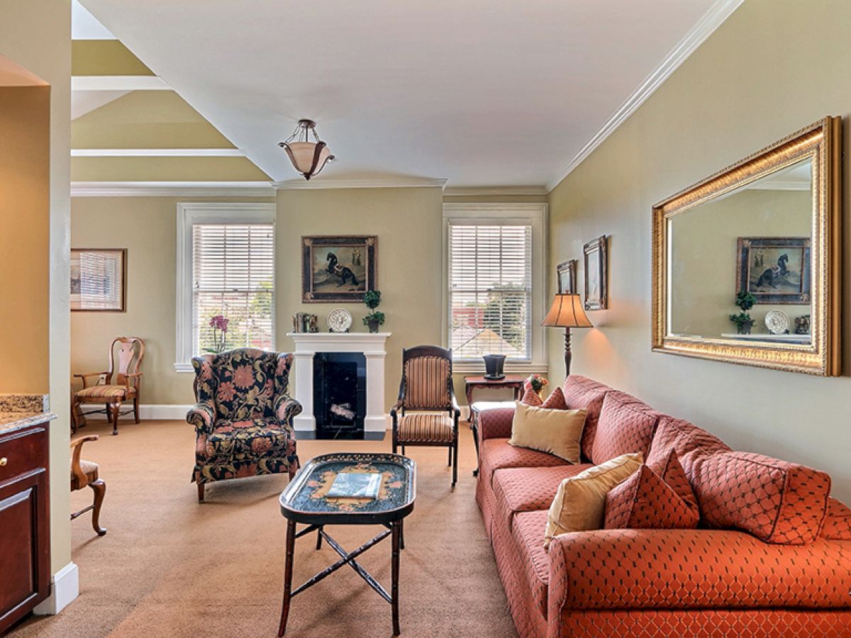 A cozy living room with a red sofa, patterned armchair, mirror, and wall art. There's a coffee table, lamp, and large window with shutters.