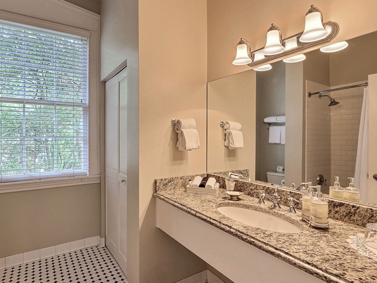 This image shows a bathroom with a granite countertop, dual sinks, a large mirror, light fixtures, a window with blinds, and a shower area.