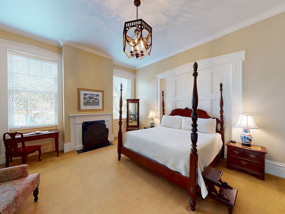 A cozy bedroom with a large four-poster bed, nightstands with lamps, a decorative fireplace, a vanity, and a comfortable chair, with ample natural light.