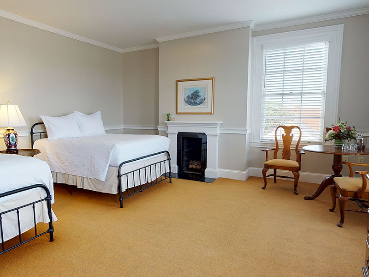 A cozy bedroom with two double beds, a small fireplace, two chairs, a round table, a chest of drawers, and a window with blinds.