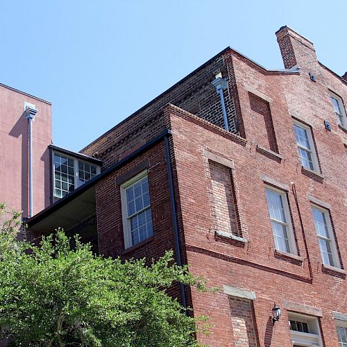 The image shows a red-brick building with multiple windows, partially surrounded by green trees, under a clear blue sky.