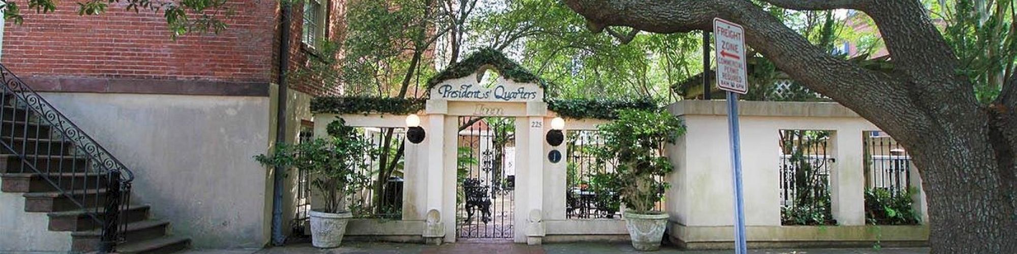 A brick pathway leads to a gated garden entrance under a large tree. The sign reads 