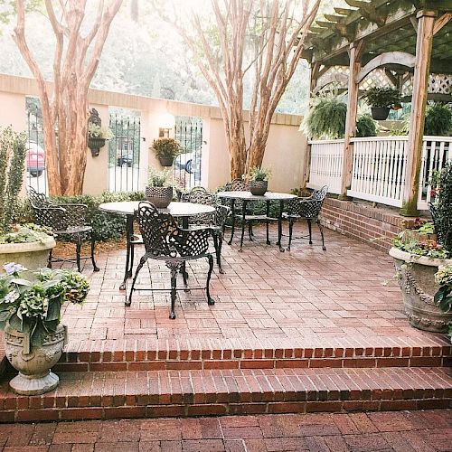 This image shows an outdoor patio area with brick flooring, wrought-iron chairs and tables, potted plants, and a wooden pergola in the background.