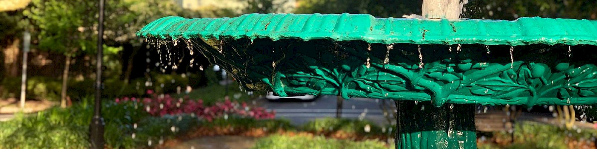 A green ornamental fountain sprays water in a park with lush trees and a red-brick path in the background, illuminated by sunlight.