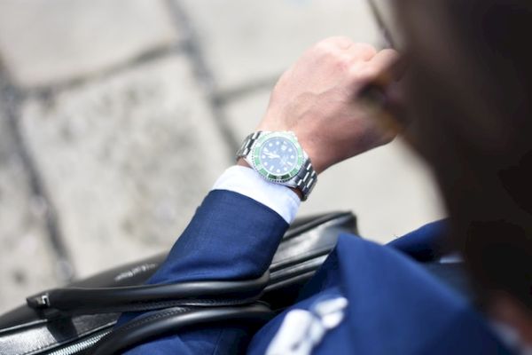 A person in a blue suit glances at a luxury watch on their wrist, holding a black bag. The scene appears to be outdoors on a stone pavement.