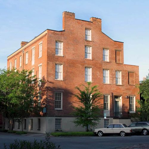 A four-story brick building with multiple windows, trees around it, and parked cars in front, set against a clear sky, on a sunny day.